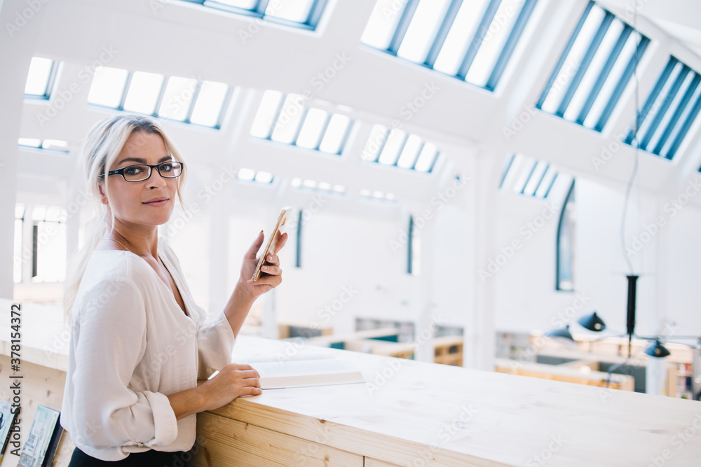 Half length portrait of beautiful female entrepreneur holding digital smartphone posing in modern workspace, charming woman in trendy glasses looking at camera during wireless browsing on cellular