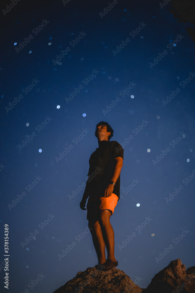 Young spanish man wearing an orange shorts shirt standing on the rocks while watching the stars in Mallorca (Spain)