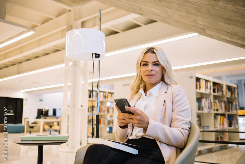 Caucasian smart casual woman holding cellphone technology and looking at camera, portrait of confident female student with modern smartphone device posing near table with education literature