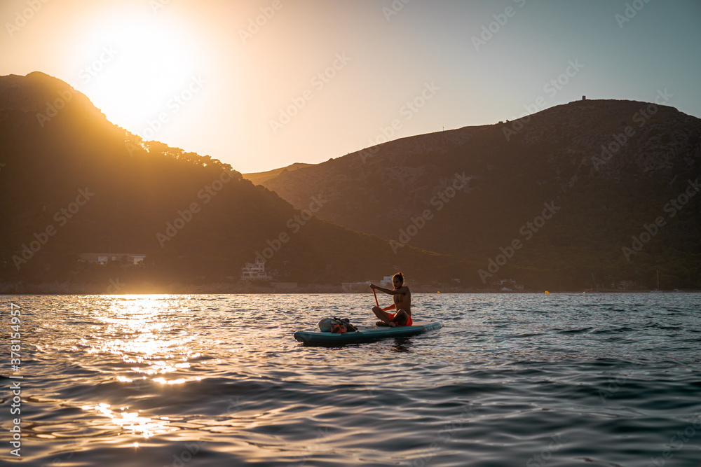 Young spanish man using his SUP on an idyllic place in Formentor, Mallorca (Spain) during sunset and sitting on it
