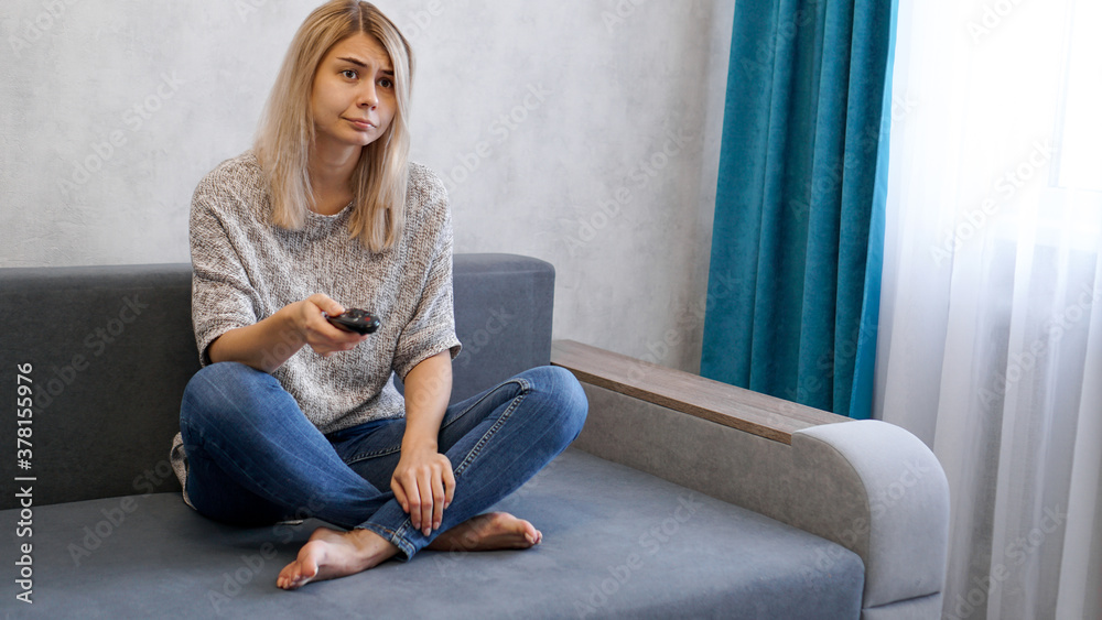Obraz premium Young woman changing TV channels with remote control sitting on sofa at home