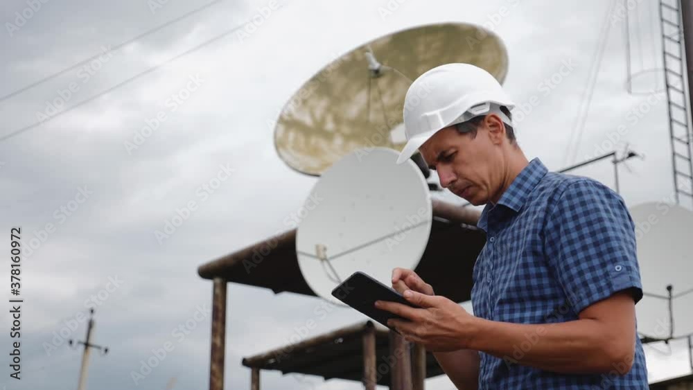 Helmet engineer and with a tablet standing on the roof of the building working with a satellite dish. Successful quality performance of tasks. Helmet engineer and with a tablet standing on the roof of the building working with a satellite dish. Successful quality performance of tasks.