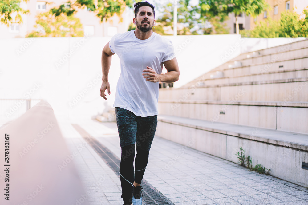 Strength Caucasian male in active wear jogging at urban city street, sportsman in earphones listening playlist songs while doing cardio training keeping healthy lifestyle and perfect body shape