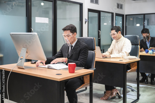 Asian employee wearing face shield working on wooden desk with desktop in new normal office