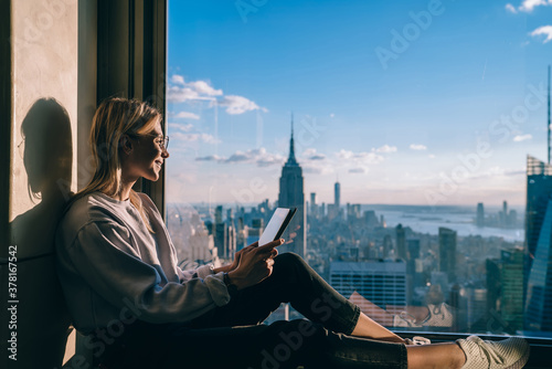 Side view of happy millennial digital nomad with modern touch pad looking in lookout window and dreaming about New York sightseeing, carefree hipster girl enjoying time in high rise building