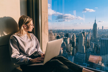 Caucasian woman in trendy clothing looking in window on metropolitan downtown admire beauty of above New York, millennial female blogger with laptop computer thinking on content idea for web sharing
