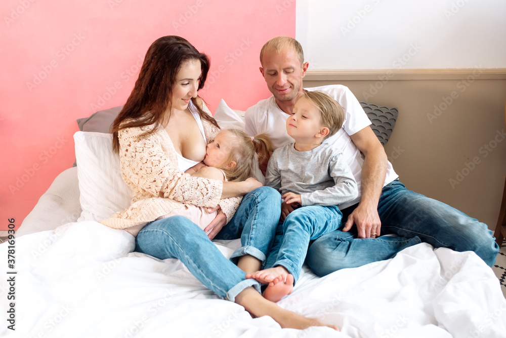 Obraz premium Beautiful young family: mom, dad, daughter and son are sitting on the bed at home in a bright bedroom and hugging 