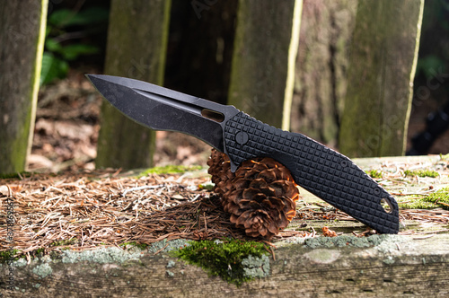 A folding knife with a black handle and a black blade. Knife and spruce cone. photo