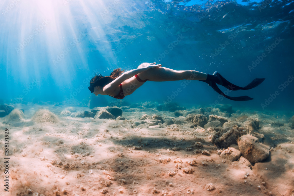 Freediver woman in bikini glides in blue sea and sun rays. Freediving with fins underwater in ocean