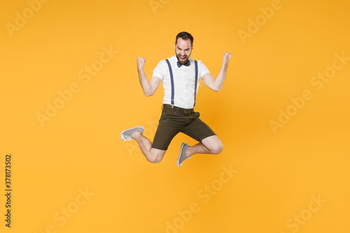 Full length portrait of happy young bearded man 20s wearing white shirt suspender shorts posing jumping doing winner gesture looking camera isolated on bright yellow color wall background studio.