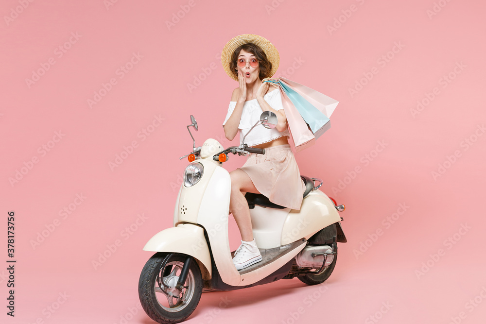 Full length portrait of amazed young woman in white clothes hat glasses hold package bag with purchases after shopping put hand on cheek driving moped isolated on pastel pink colour background studio. Full length portrait of amazed young woman in white clothes hat glasses hold package bag with purchases after shopping put hand on cheek driving moped isolated on pastel pink colour background studio.
