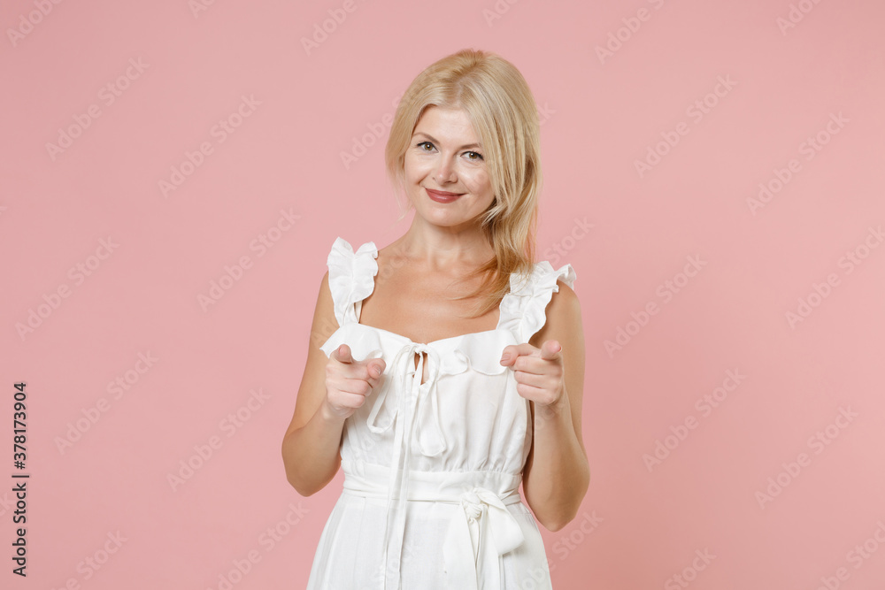 Smiling cheerful cute stunning beautiful attractive young blonde woman wearing white summer dress standing pointing index fingers on camera isolated on pastel pink colour background, studio portrait. Smiling cheerful cute stunning beautiful attractive young blonde woman wearing white summer dress standing pointing index fingers on camera isolated on pastel pink colour background, studio portrait.