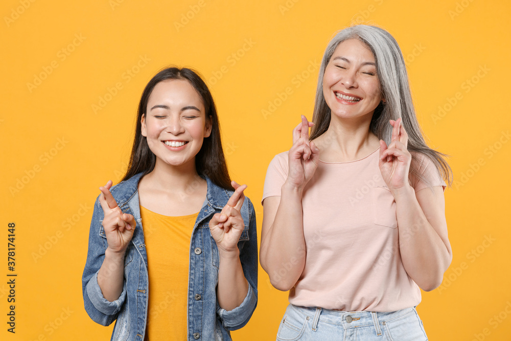 Smiling family two asian women girls mother daughter in casual clothes waiting for special moment keeping fingers crossed eyes closed making wish isolated on yellow color background studio portrait. Smiling family two asian women girls mother daughter in casual clothes waiting for special moment keeping fingers crossed eyes closed making wish isolated on yellow color background studio portrait.
