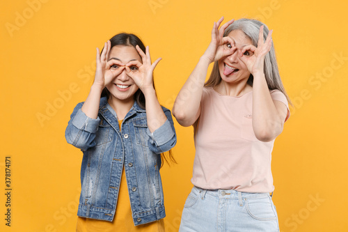 Funny family asian women girls gray-haired mother brunette daughter in casual clothes posing hold hands near eyes, imitating glasses or binoculars isolated on yellow color background studio portrait.