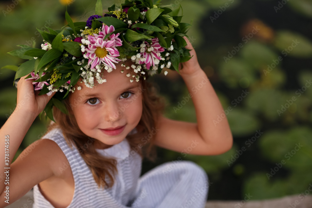 Fototapeta premium Cute little girl wearing wreath made of beautiful flowers near pond