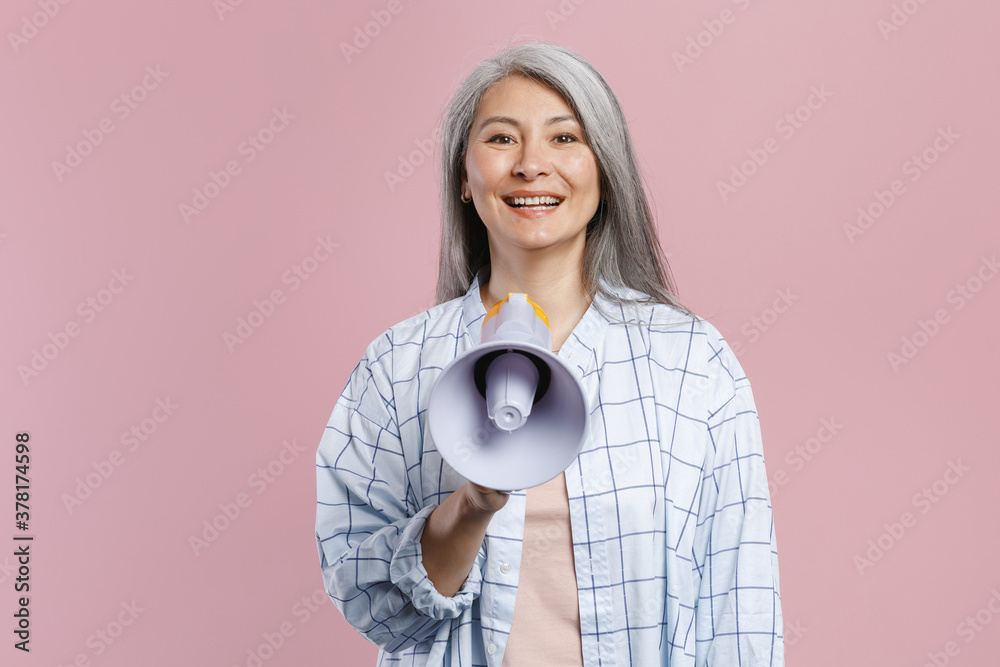 Smiling cheerful gray-haired asian woman wearing basic white checkered shirt standing hold in hand screaming in megaphone looking camera isolated on pastel pink colour background, studio portrait. Smiling cheerful gray-haired asian woman wearing basic white checkered shirt standing hold in hand screaming in megaphone looking camera isolated on pastel pink colour background, studio portrait.