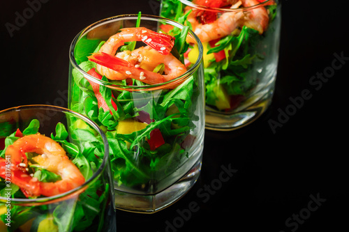 A salad of mango, sweet pepper, rukkola and garlic-fried shrimp in a glass. Close up. Selective focus. Black background.