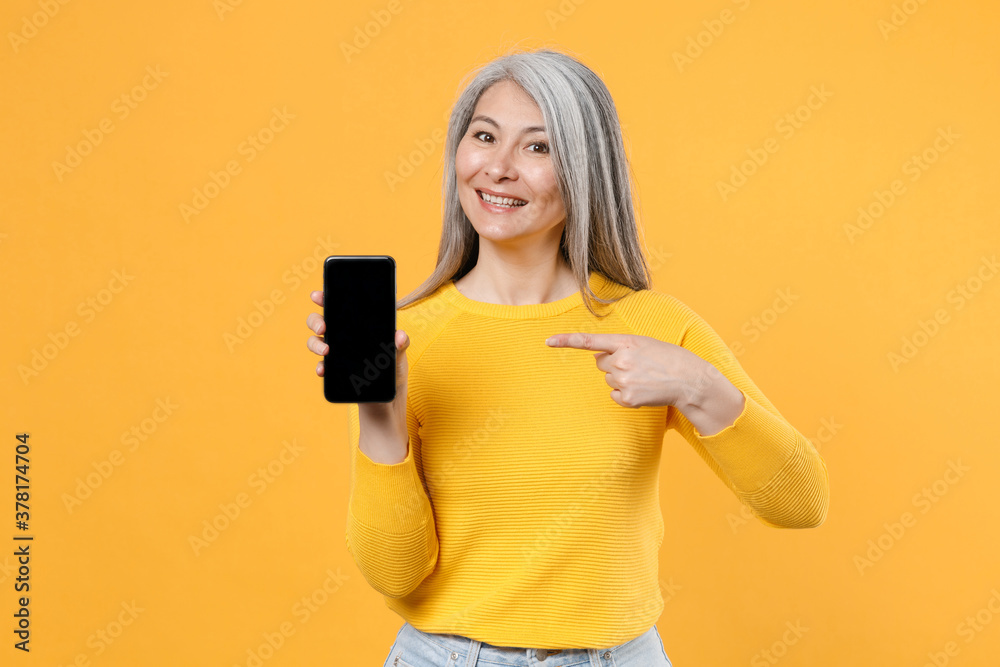 Smiling gray-haired asian woman in casual clothes pointing index finger on mobile cell phone with blank empty screen mock up copy space isolated on bright yellow colour background, studio portrait. Smiling gray-haired asian woman in casual clothes pointing index finger on mobile cell phone with blank empty screen mock up copy space isolated on bright yellow colour background, studio portrait.