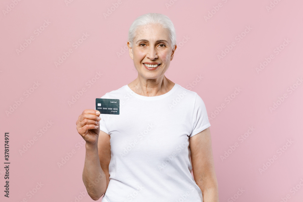 Smiling elderly gray-haired female woman 60s 70s wearing white casual blank design t-shirt posing hold credit bank card looking camera isolated on pastel pink color wall background studio portrait. Smiling elderly gray-haired female woman 60s 70s wearing white casual blank design t-shirt posing hold credit bank card looking camera isolated on pastel pink color wall background studio portrait.
