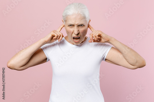 Crazy angry elderly gray-haired female woman wearing white blank casual t-shirt posing covering ears with fingers looking camera screaming isolated on pastel pink color background studio portrait.