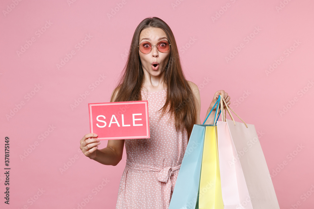 Fototapeta premium Shocked young brunette woman in pink summer dotted dress glasses hold package bag with purchases after shopping sign with SALE title looking camera isolated on pastel pink background studio portrait.