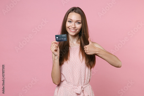 Smiling young brunette woman 20s wearing pink summer dotted dress posing holding in hand credit bank card showing thumb up looking camera isolated on pastel pink color wall background studio portrait.