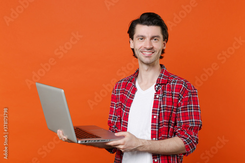 Smiling handsome young brunet man 20s wearing white t-shirt red checkered shirt posing standing working on laptop pc computer looking camera isolated on orange color wall background studio portrait.