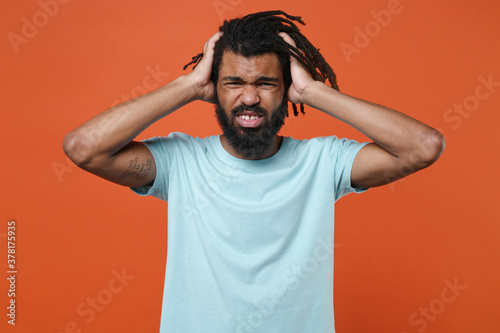 Tired preoccupied young african american man guy wearing blue casual t-shirt posing isolated on bright orange background studio portrait. People lifestyle concept. Put hands on head, having headache.