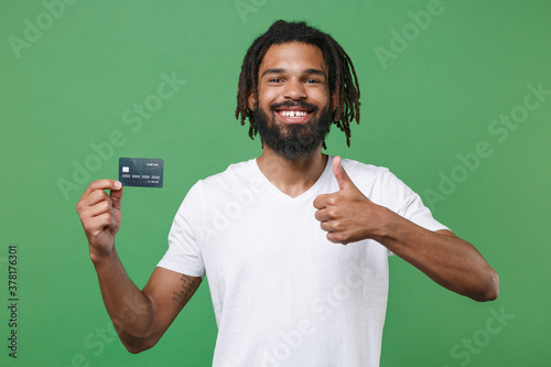 Smiling funny young african american man guy with dreadlocks 20s wearing white casual t-shirt posing hold credit bank card showing thumb up isolated on green color wall background studio portrait.