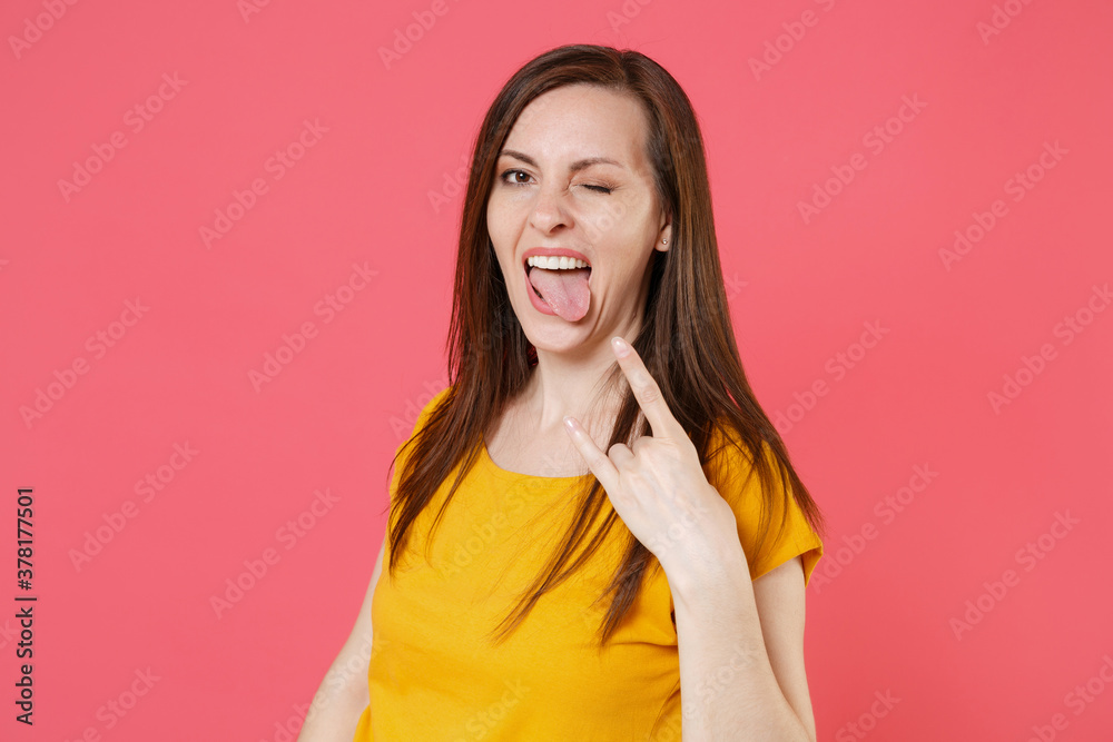 Blinking funny young brunette woman 20s wearing yellow casual t-shirt posing standing depicting heavy metal rock sign horns up gesture showing tongue isolated on pink color background studio portrait. Blinking funny young brunette woman 20s wearing yellow casual t-shirt posing standing depicting heavy metal rock sign horns up gesture showing tongue isolated on pink color background studio portrait.