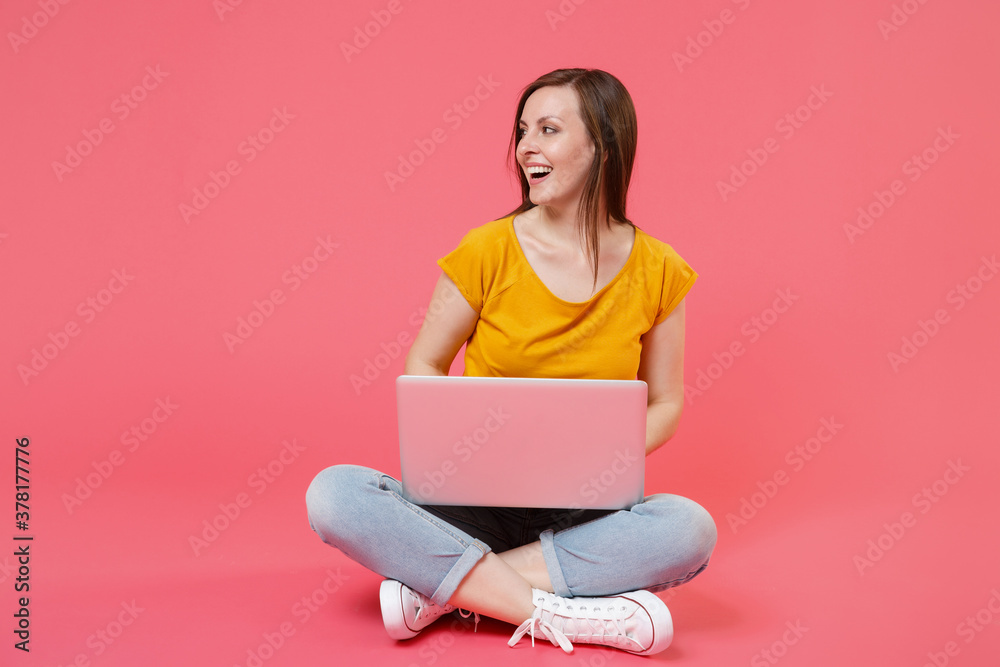 Full length portrait of cheerful smiling young brunette woman 20s wearing yellow t-shirt sitting on floor working on laptop pc computer looking aside isolated on pink color wall background studio. Full length portrait of cheerful smiling young brunette woman 20s wearing yellow t-shirt sitting on floor working on laptop pc computer looking aside isolated on pink color wall background studio.