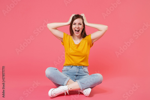 Full length portrait of cheerful funny excited beautiful attractive young brunette woman 20s wearing yellow t-shirt sitting on floor put hands on head isolated on pink color wall background studio. © ViDi Studio