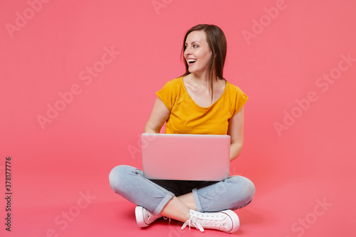 Full length portrait of cheerful smiling young brunette woman 20s wearing yellow t-shirt sitting on floor working on laptop pc computer looking aside isolated on pink color wall background studio.