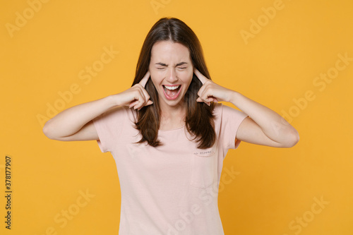 Crazy frustrated young brunette woman in pastel pink casual t-shirt standing posing covering ears with fingers keeping eyes closed screaming isolated on bright yellow color background studio portrait.