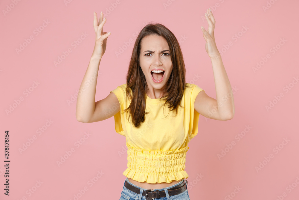 Angry irritated young brunette woman 20s wearing yellow casual t-shirt posing standing spreading hands swearing screaming looking camera isolated on pastel pink color wall background studio portrait. Angry irritated young brunette woman 20s wearing yellow casual t-shirt posing standing spreading hands swearing screaming looking camera isolated on pastel pink color wall background studio portrait.