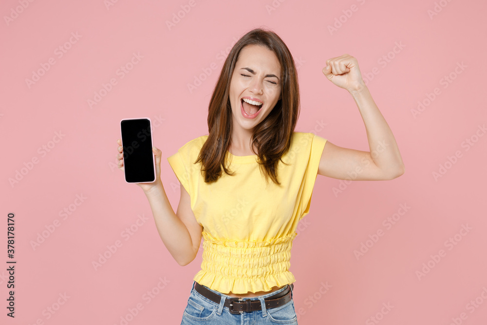 Joyful young brunette woman 20s wearing yellow casual t-shirt hold mobile phone with blank empty screen mock up copy space doing winner gesture isolated on pastel pink wall background studio portrait. Joyful young brunette woman 20s wearing yellow casual t-shirt hold mobile phone with blank empty screen mock up copy space doing winner gesture isolated on pastel pink wall background studio portrait.