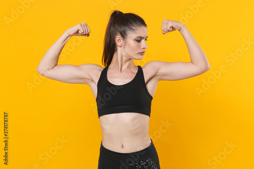 Strong attractive young fitness sporty woman 20s wearing black sportswear posing working out training showing biceps, muscles looking aside isolated on bright yellow color background studio portrait.