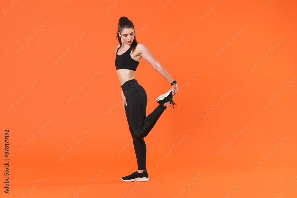 Full length side view portrait young fitness sporty woman in black sportswear posing training working out doing stretching exercising for legs looking down isolated on orange color background studio.