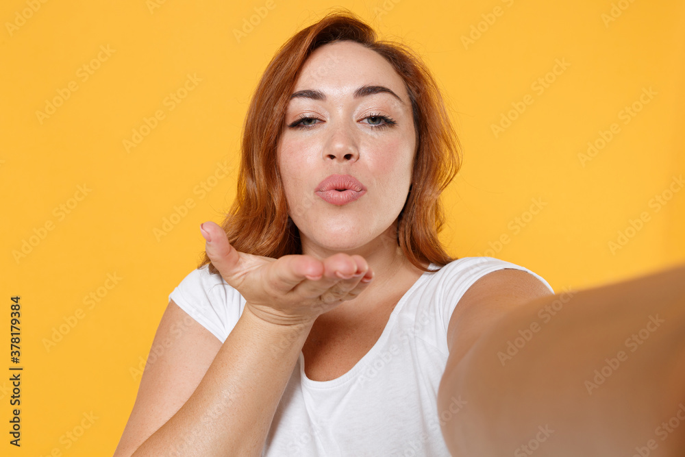 Close up pretty young redhead plus size body positive female woman 20s in white t-shirt doing selfie shot on mobile phone blowing sending air kiss isolated on yellow color background studio portrait. Close up pretty young redhead plus size body positive female woman 20s in white t-shirt doing selfie shot on mobile phone blowing sending air kiss isolated on yellow color background studio portrait.