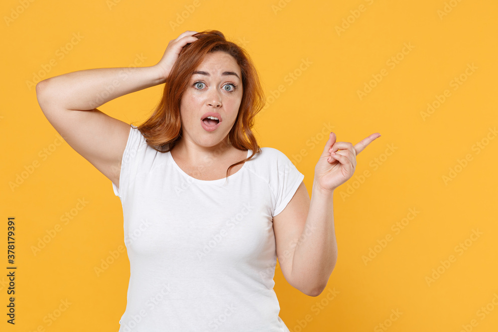 Shocked young redhead plus size body positive female woman in white casual t-shirt put hand on head point index finger aside on mock up copy space isolated on yellow color background studio portrait. Shocked young redhead plus size body positive female woman in white casual t-shirt put hand on head point index finger aside on mock up copy space isolated on yellow color background studio portrait.
