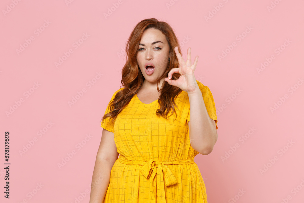 Stunning attractive shocked young redhead plus size body positive female woman girl in yellow dress posing showing OK gesture looking camera isolated on pastel pink color background studio portrait. Stunning attractive shocked young redhead plus size body positive female woman girl in yellow dress posing showing OK gesture looking camera isolated on pastel pink color background studio portrait.