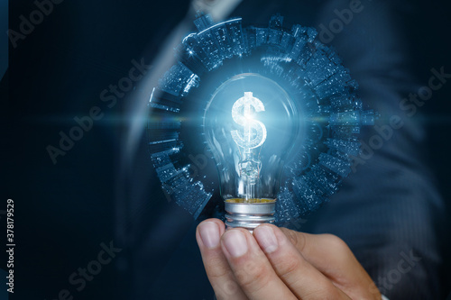Businessman showing light bulb with burning dollar hologram inside on blue background.