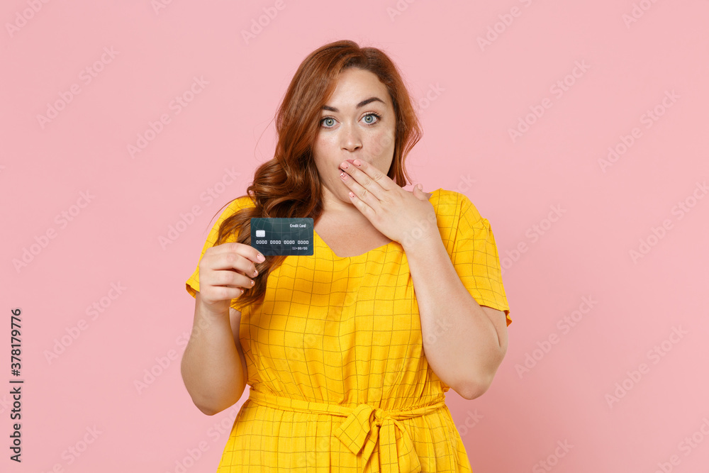 Shocked young redhead plus size body positive female woman girl 20s in yellow dress posing hold credit bank card covering mouth with hand isolated on pastel pink color wall background studio portrait. Shocked young redhead plus size body positive female woman girl 20s in yellow dress posing hold credit bank card covering mouth with hand isolated on pastel pink color wall background studio portrait.