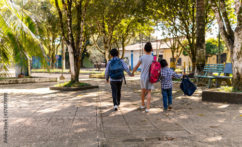 three children walking hand in hand in a park with school bag