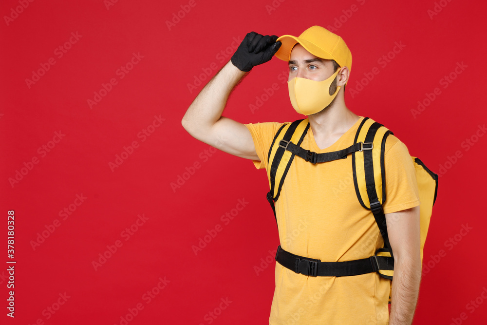 Delivery employee man in yellow cap face mask gloves t-shirt uniform thermal bag backpack with food work courier service during quarantine coronavirus covid-19 virus isolated on red background studio.