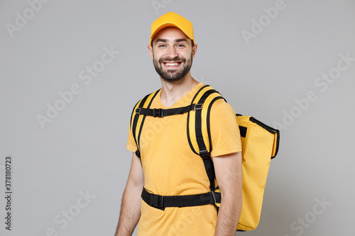 Delivery employee man guy male 20s in yellow cap t-shirt uniform thermal food bag backpack work courier service during quarantine coronavirus covid-19 virus, posing isolated on gray background studio.