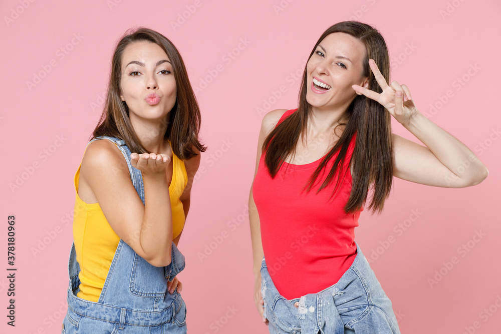 Two pretty cheerful young women friends 20s wearing casual denim clothes posing blowing sending air kiss showing victory sign looking camera isolated on pastel pink colour background, studio portrait. Two pretty cheerful young women friends 20s wearing casual denim clothes posing blowing sending air kiss showing victory sign looking camera isolated on pastel pink colour background, studio portrait.