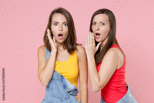 Two shocked worried young women friends 20s in casual denim clothes posing whispers gossip and tells secret with hand gesture looking camera isolated on pastel pink colour background, studio portrait.