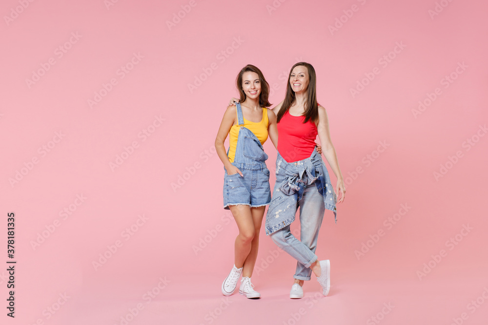 Full length portrait of two smiling cheerful attractive young brunette women friends 20s wearing casual denim clothes posing hugging looking camera isolated on pastel pink colour background in studio. Full length portrait of two smiling cheerful attractive young brunette women friends 20s wearing casual denim clothes posing hugging looking camera isolated on pastel pink colour background in studio.
