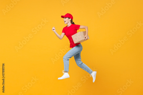 Full length body jumping delivery employee woman in red cap blank t-shirt uniform work courier in service during quarantine coronavirus covid-19 virus hold cardboard box isolated on yellow background. © ViDi Studio